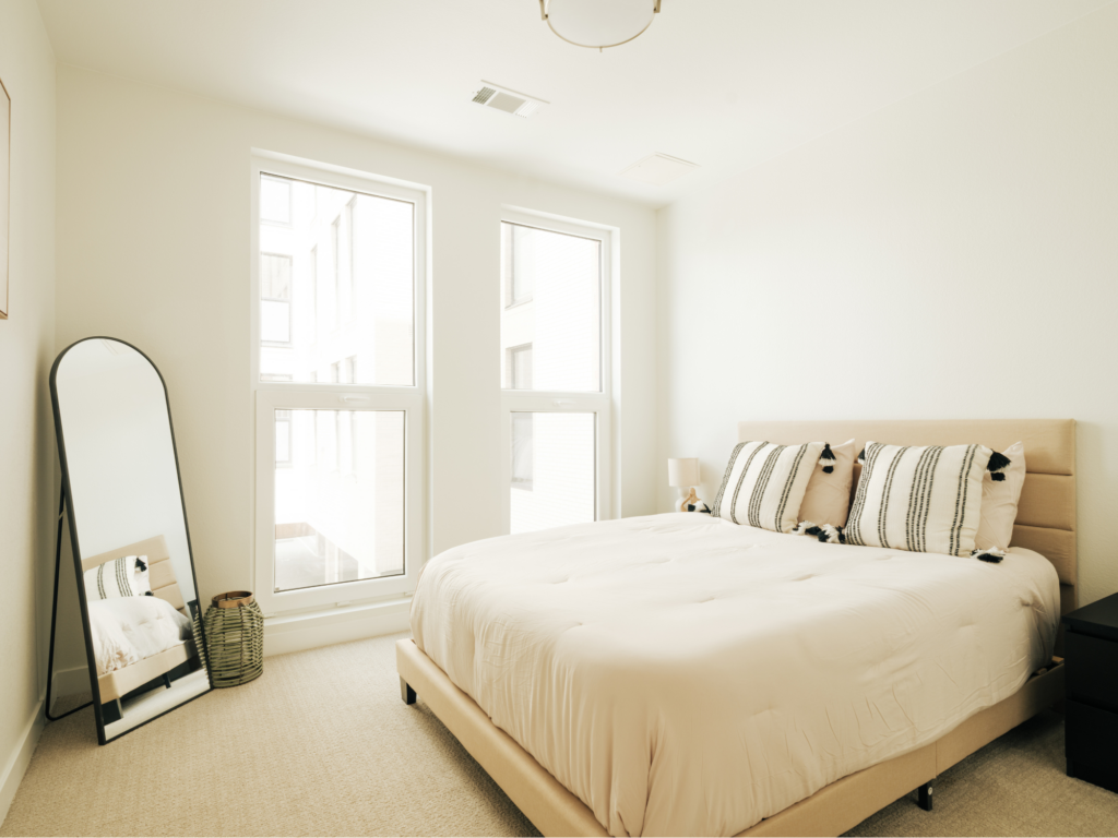 Bright minimalist bedroom with upholstered bed, striped pillows, full-length mirror, and two large windows.