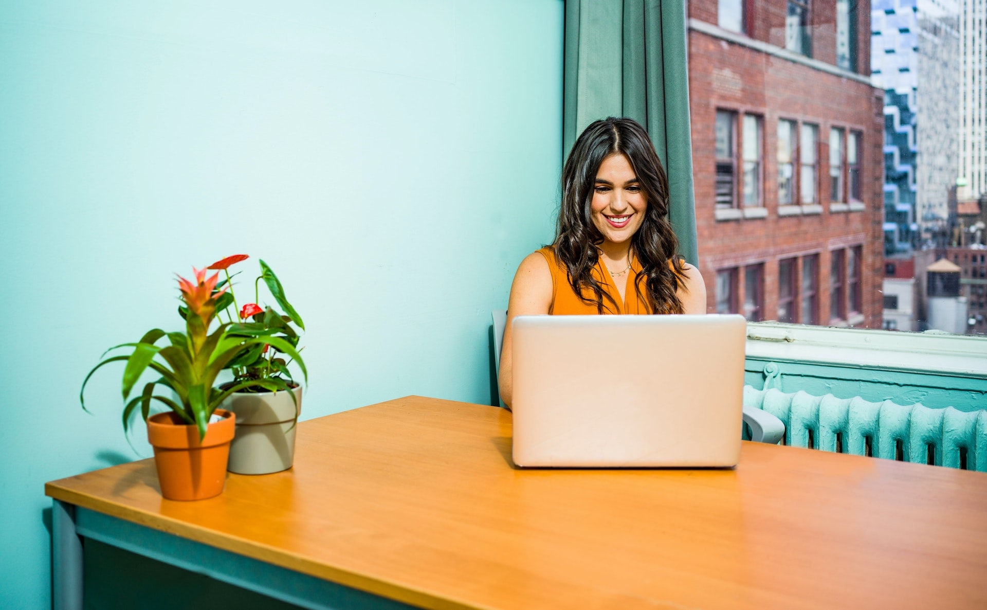 Smiling woman in orange top working on laptop at wooden desk with potted plants, city view through window.