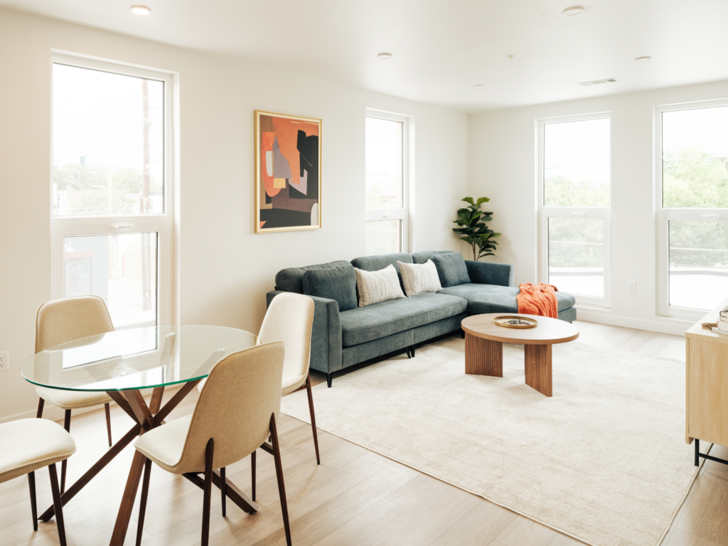 Bright modern living room with gray sectional sofa, round wooden coffee table, abstract wall art, and glass dining table with four chairs near large windows.