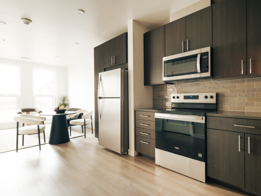 Modern apartment kitchen with dark wood cabinets, stainless steel appliances, and adjacent dining area with round table and white chairs.